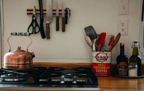 A kitchen countertop with a copper kettle, knives, utensils, and bottles, featuring a can of Colombian coffee.