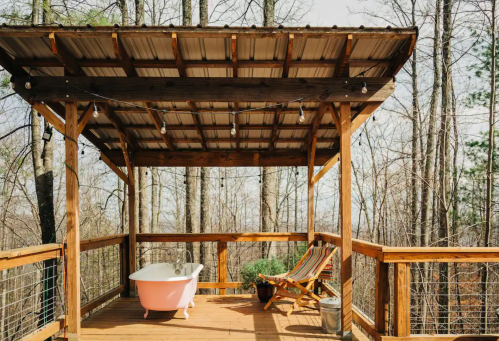 A wooden deck with a pink bathtub and a chair, surrounded by trees and string lights overhead.