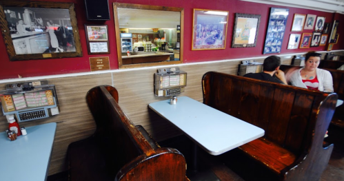 A cozy diner interior with wooden booths, a blue table, and vintage decor on the walls. Two patrons are seated.