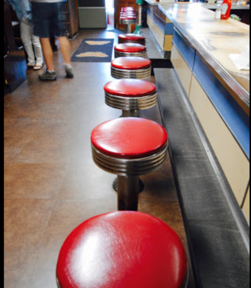 A row of red vinyl stools at a diner counter, with a wooden bar and a glimpse of patrons in the background.
