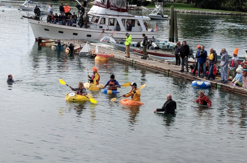 A group of kayakers in colorful kayaks paddles near a dock with spectators watching from the shore.