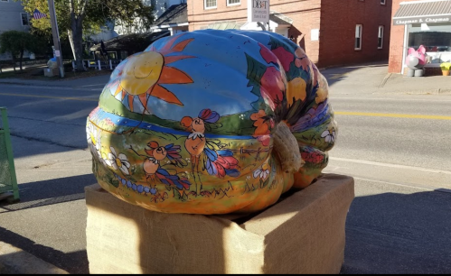 A large, painted pumpkin sculpture with colorful flowers and chickens, displayed on a wooden platform in a town setting.