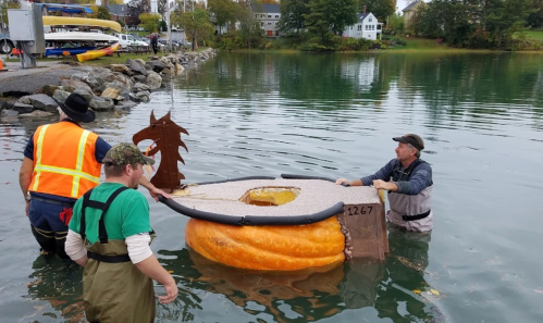 Three people prepare a large pumpkin boat on a calm water surface, with a scenic backdrop of houses and trees.