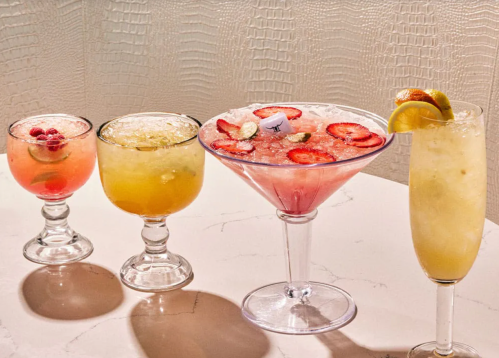 Four colorful cocktails with fruit garnishes on a marble table against a textured background.