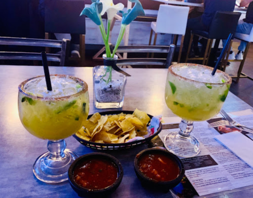 Two large cocktails with lime and mint, served with tortilla chips and salsa, alongside a small vase of flowers.