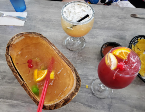 Three colorful cocktails on a table: a fruity drink in a wooden bowl, a margarita, and a red cocktail with fruit garnish.
