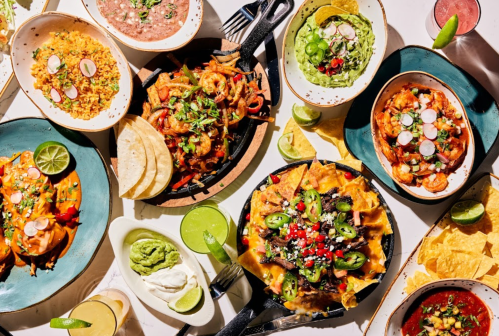 A vibrant spread of Mexican dishes, including nachos, tacos, guacamole, and rice, garnished with fresh herbs and lime.