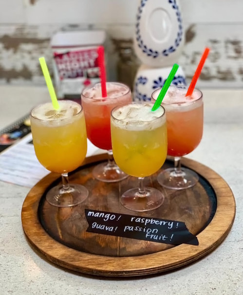 Four colorful cocktails on a wooden tray, labeled mango, raspberry, guava, and passion fruit, with straws.