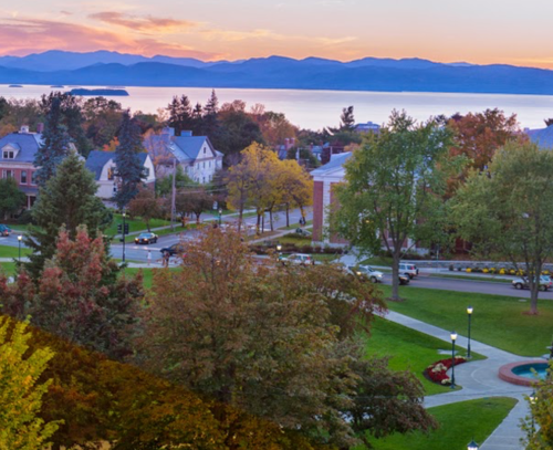 A scenic view of a town with trees in autumn colors, mountains in the background, and a calm lake at sunset.