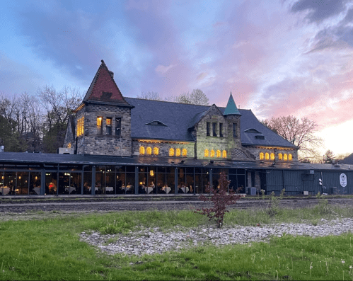 Historic stone building with large windows, surrounded by greenery, under a colorful sunset sky.