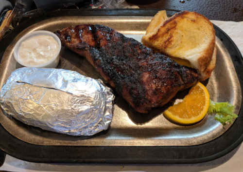 A grilled steak on a metal plate with a baked potato, toast, an orange slice, and a small cup of sauce.