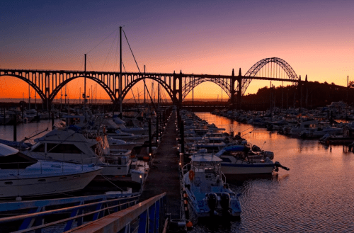Sunset over a marina with boats, featuring a bridge in the background and vibrant orange and purple sky.