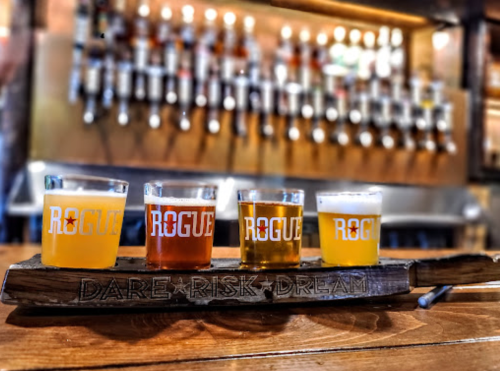 Four glasses of beer on a wooden flight paddle, labeled "Rogue," with a brewery tap wall in the background.