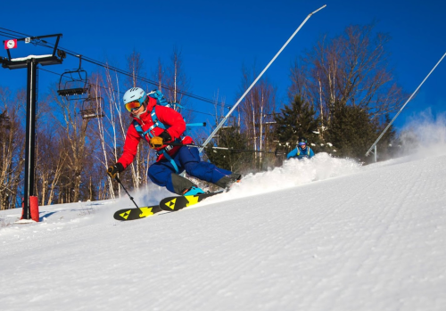 A skier in a red jacket glides down a snowy slope with a ski lift in the background under a clear blue sky.