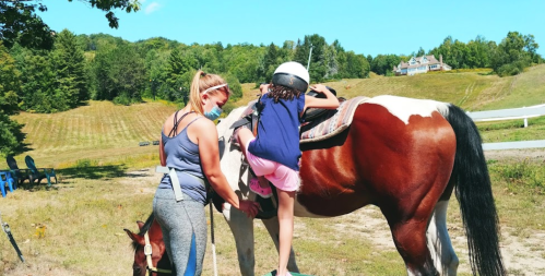 A child in a helmet climbs onto a brown and white horse with the help of an adult in a sunny outdoor setting.