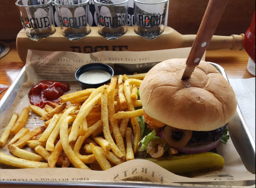 A burger with a wooden skewer, served with fries, ketchup, ranch dressing, and a pickle on a tray.