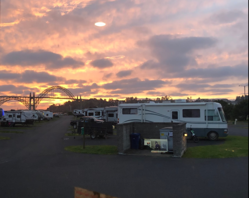 A scenic view of an RV park at sunset, with a bridge in the background and colorful clouds in the sky.