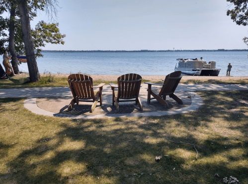 Three wooden chairs facing a calm lake, with a boat nearby and trees casting shadows on the grass.