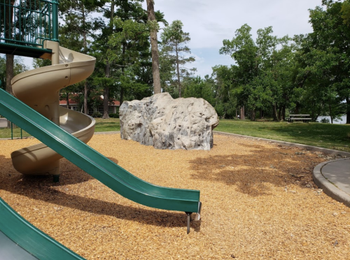 A playground featuring a green slide, a large rock formation, and wood chip ground cover surrounded by trees.