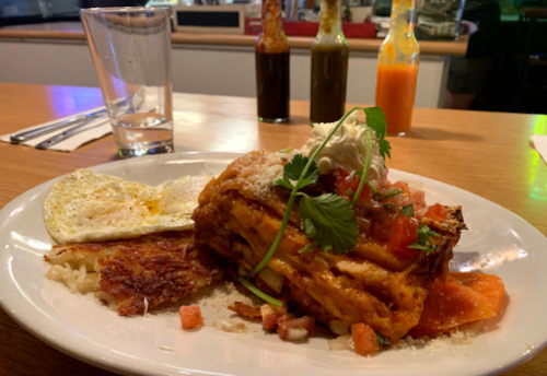 A plate of enchiladas topped with sour cream, tomatoes, and cilantro, served with eggs and hash browns in a restaurant setting.