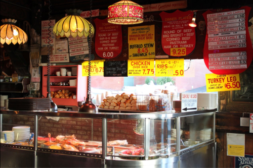 A vintage deli display with menu signs, showcasing sandwiches, soups, and meats under warm lighting.
