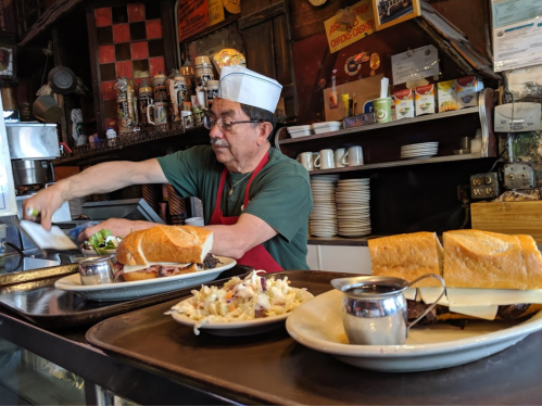 A chef prepares sandwiches at a counter, with plates of food and condiments in a cozy restaurant setting.
