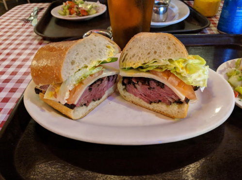 A sandwich with layers of meat, lettuce, and tomato, served on a plate with a side salad and a drink in the background.