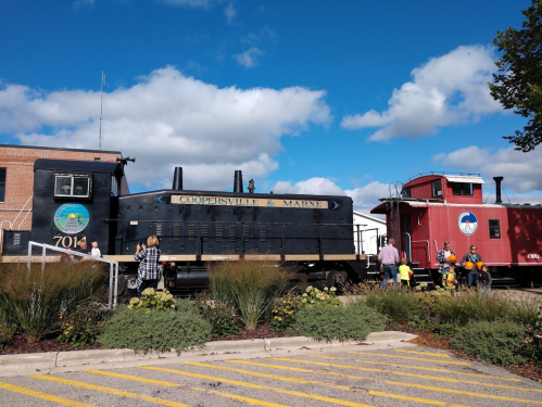 A black locomotive and a red caboose at a train station, with people exploring the area under a blue sky.