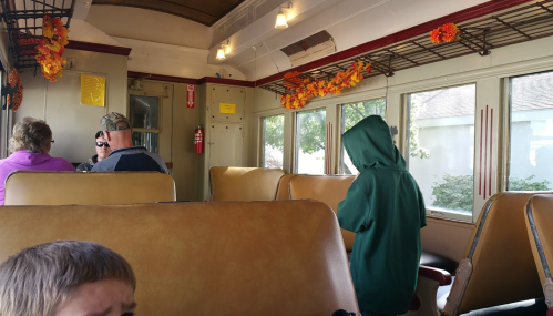 Interior of a train car with passengers seated, a child in a green hoodie stands near the window. Fall decorations are visible.