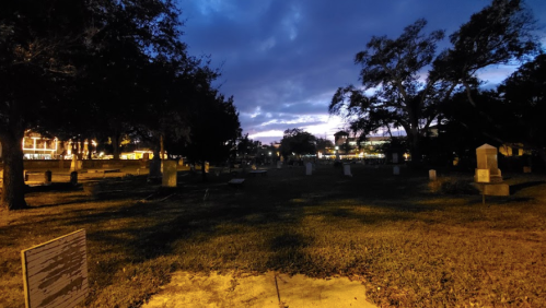 A dimly lit cemetery at dusk, with gravestones and trees silhouetted against a cloudy sky.