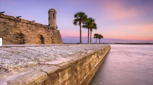 A stone wall with a tower beside a calm waterway, framed by palm trees under a colorful sunset sky.