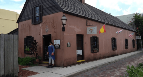 Historic building with a pink exterior, flags, and a person entering. Cobblestone path in front.