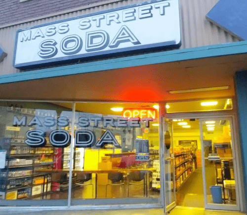 Storefront of Mass Street Soda with large windows, "OPEN" sign, and shelves of beverages visible inside.