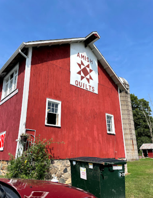 Red barn with a quilt pattern sign, surrounded by greenery and a dumpster, under a clear blue sky.