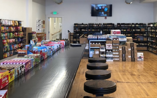 Interior of a store with shelves of snacks and beverages, a long counter, and a TV displaying information.