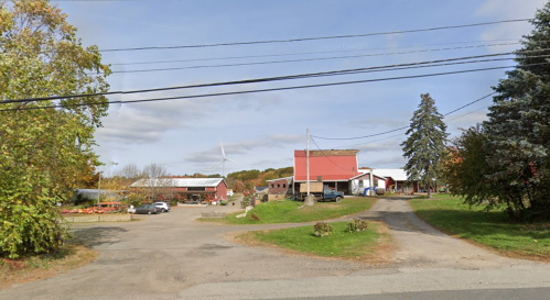 A rural scene featuring a farm with a red-roofed barn, wind turbine, and trees under a partly cloudy sky.