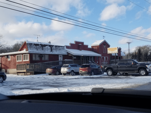 A red barn-style building with snow-covered ground and parked cars in front, under a partly cloudy sky.