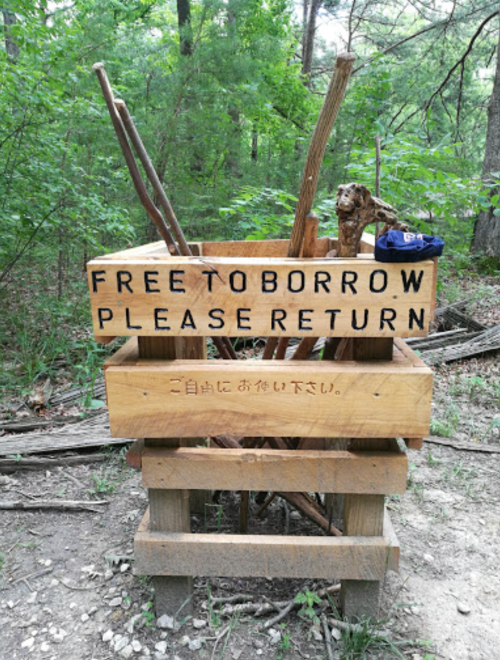 A wooden borrowing station in a forest, with a sign that reads "FREE TO BORROW PLEASE RETURN" in English and Japanese.