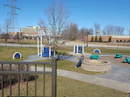 A playground with blue equipment, a tree, and a seating area, set against a clear blue sky and distant buildings.