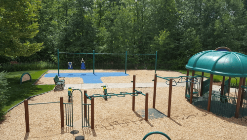 A playground with swings, climbing structures, and a playhouse, surrounded by trees and gravel ground.
