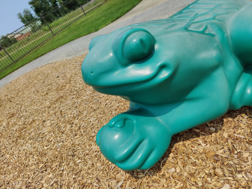 A large, teal frog sculpture sits on a playground surface covered with wood chips.