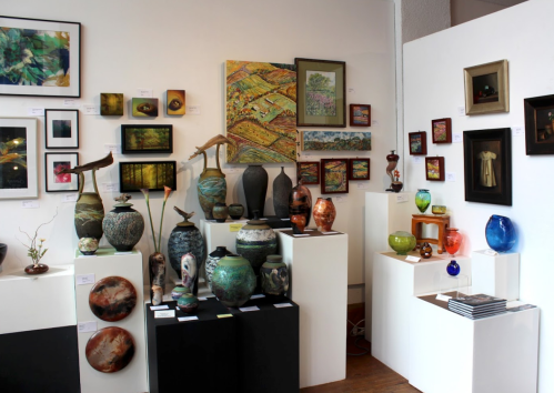 Art gallery display featuring various pottery, paintings, and sculptures on white pedestals and walls.