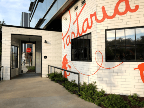 Exterior of a restaurant named "Pastaria," featuring a white brick wall with bold orange lettering and a welcoming entrance.