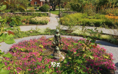 A garden with vibrant flowers surrounding a central statue of a child holding a globe, set in a lush landscape.