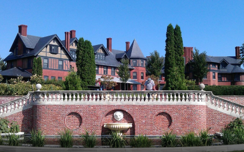 A grand brick mansion with multiple gables, surrounded by lush greenery and a decorative fountain in the foreground.