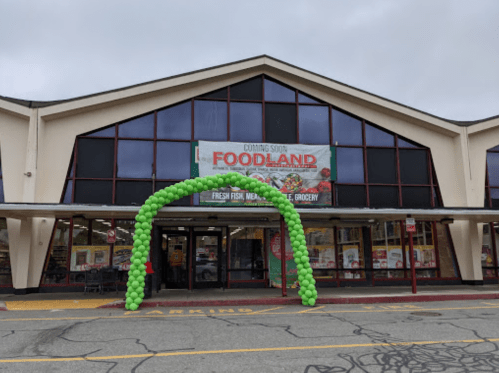A storefront with a large "Coming Soon Foodland" banner and a green balloon arch at the entrance.