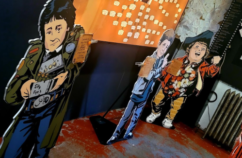 Three life-sized cardboard cutouts of characters in colorful outfits, holding signs, set against a textured wall.