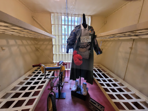 A mannequin dressed in a costume stands in a small, empty room with a bicycle nearby, resembling a jail cell.