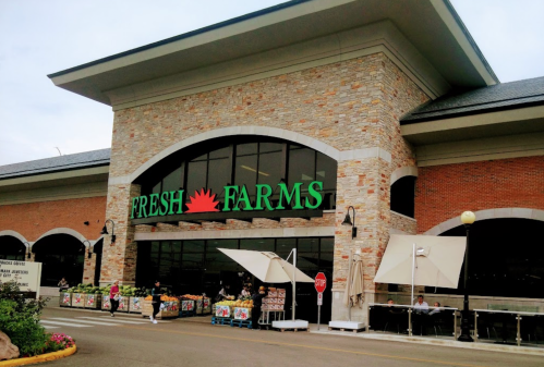 Exterior of Fresh Farms grocery store with a stone facade and large windows, featuring outdoor seating and displays.