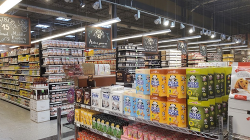 A grocery store aisle filled with various food products, including snacks and sauces, under bright lighting.